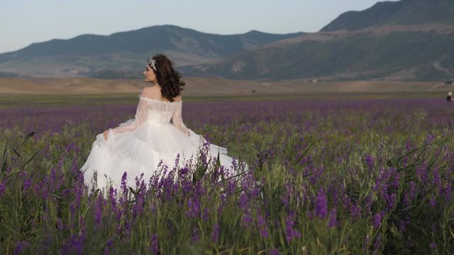 cheerful bride in elegant wedding dress with bouffant skirt in blooming purple flowers against mountains slow motion