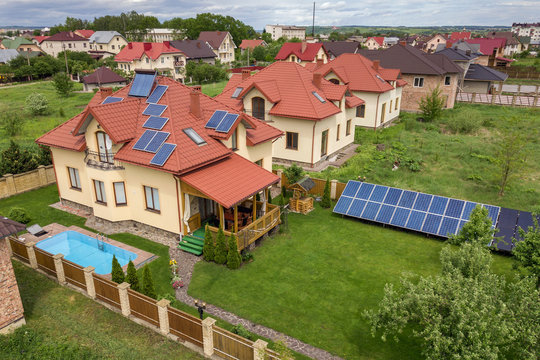 Aerial View Of A New Autonomous House With Solar Panels And Water Heating Radiators On The Roof And Green Yard With Blue Swimming Pool.