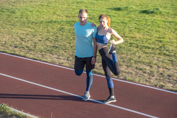 Obraz premium Young couple of fit sportsmen boy and girl doing warming up exercises before running on red tracks of public stadium outdoors.