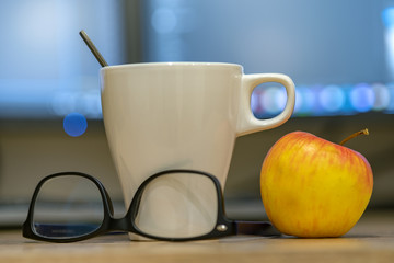White cup of coffee or tea, glasses and a red apple on blurred background of personal computer screen.