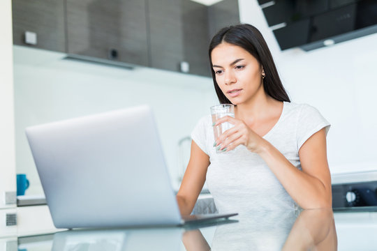 Young Woman sitting in kitchen table, looking at laptop computer screen while drink water from glass