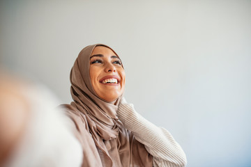 Happy young muslim woman take self portrait with handphone at home. Happy arab woman in hijab with mobile phone making selfie. Portrait of smiling girl, posing.