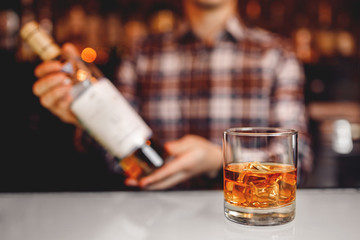 Glass of whiskey with ice stands on bar counter, dark brown background