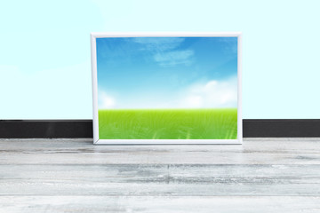 Landscape wooden frame. Wooden picture frame with an abstract countryside with cloudy sky  on a rustic table in front of light blue background. Template framing for modern art.