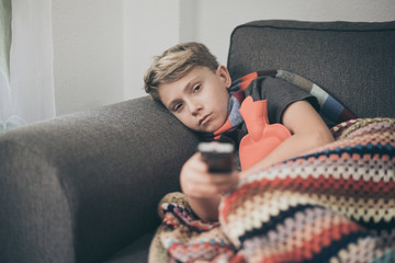 Sick boy watching tv with woolen blanket and hot water bottle. Sad teen with the flu rests at home in a cold winter day. Child with seasonal infections and fever Health disease and drugs concept