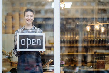 woman stands and opens a wide sign through the shop window and smile.