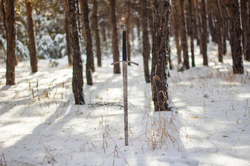 Knight's long sword in the snow against the background of a pine winter forest