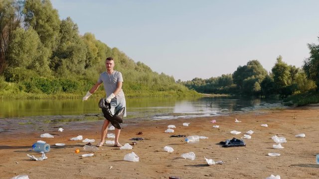 Man in gray t-shirt and blue denim shorts stands among the trash and dances the floss on banks of dry and polluted river, lake. Ecological catastrophy. Anthropogenic influence. 4K slowmotion footage.