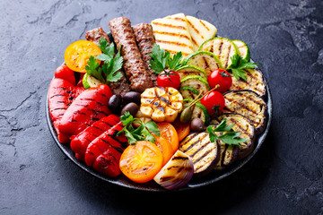 Grilled meat kebabs and vegetables on a plate. Black stone background. Close up.
