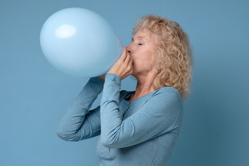Beautiful senior blonde woman preparing for party blowing up blue balloon