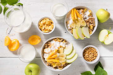 Bowls with granola, fruit, yogurt and two glasses with milk on a white wooden background. Healthy breakfast cereal