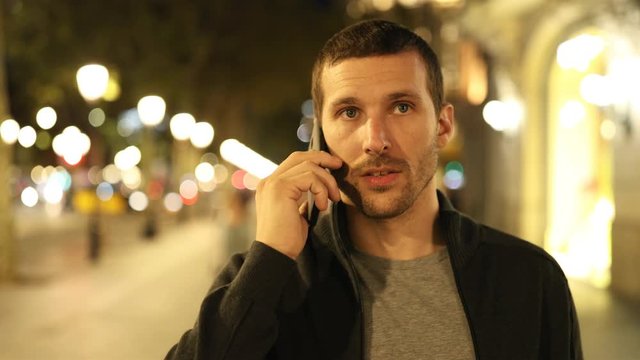 Front view of a serious man walking talking on phone in the night in the street