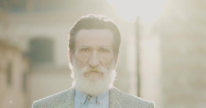 Portrait of the senior Caucasian man with grey beard looking straigt to the camera with shrewd eyes. Outdoors. Close up.