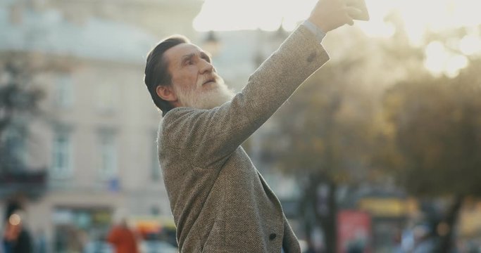 Old Caucasian man with a long beard standing in the center city and taking selfie photos on the smartphone camera. Outside.
