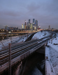 Fototapeta premium Industrial landscape with many railroad ways going far and skyscrapers with power plant pipes on horizon at night in winter period vertical view