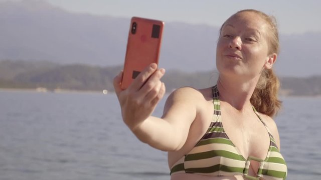 Female tourist posing when taking pictures of yourself on phone on background of sea. Adult woman makes selfie using smartphone at sea.