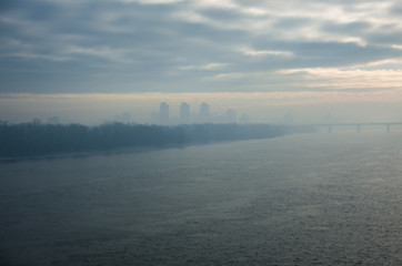 Fototapeta premium Cold early winter morning, light haze and fog rise above the calm water of the river, on the shore there are trees without leaves.Bridge and the silhouette of the buildings in the distance. Kyiv. 