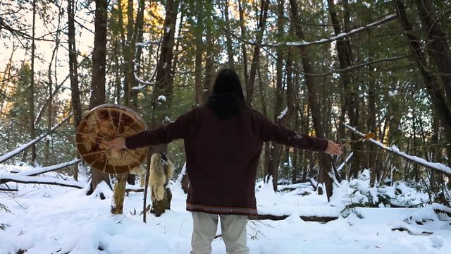 Slow motion and rear view footage of a spiritual man during in a winter forest, raising arms in air with a scared native rawhide drum and beater