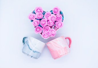 Flatlay composition. Two marble cups in pink and gray and pink flowers in a heart-shaped casket on a white background. Valentines day concept