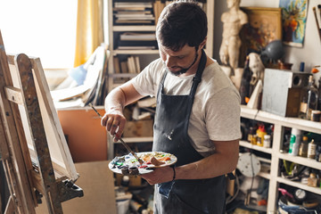 Male painter at art studio indoors with palette mixing colors ready to draw concentrated