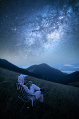 Young woman drinking coffee at mountains on the background of the milky way. Woman looking at beautiful milky way. Starry sky with hills at summer