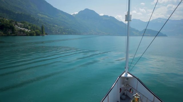 Approaching amazing mountains or lakeside town in Switzerland. Front view from ship nose pov with magnificent lake scenery. Calm smooth surface of colorful blue lake. Clear sky reflected in water.