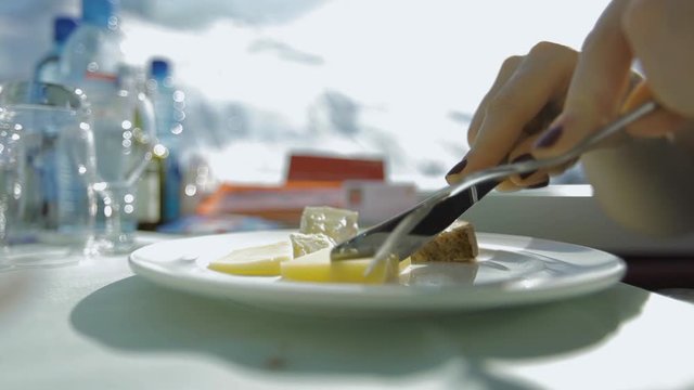 Woman's hands slices swiss cheese on a white plate in railroad restaurant by the window in the moving train. Magnificent view of winter mountains in Alps. Glacier Express train in Switzerland.