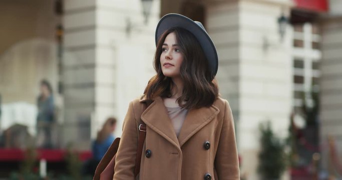 Caucasian stylish attractive young woman model or traveller in a hat and cout having a walk at the city center street and looking at the side. Outdoor.