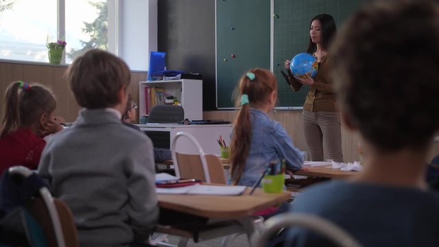 Focused elementary age schoolkids listening to asian woman teacher during geography lesson in junior school. Skillful teacher using globe while telling about geographic coordinates to curious students