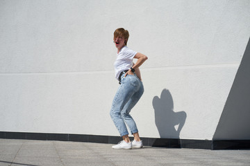Fototapeta na wymiar Shorthair redhair girl shows her butt, standing in front of white wall outdors in summer