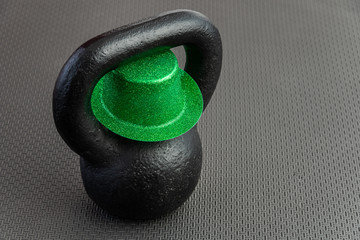 Naklejka na ściany i meble Black iron kettlebell with green leprechaun hat on a black gym floor, holiday fitness