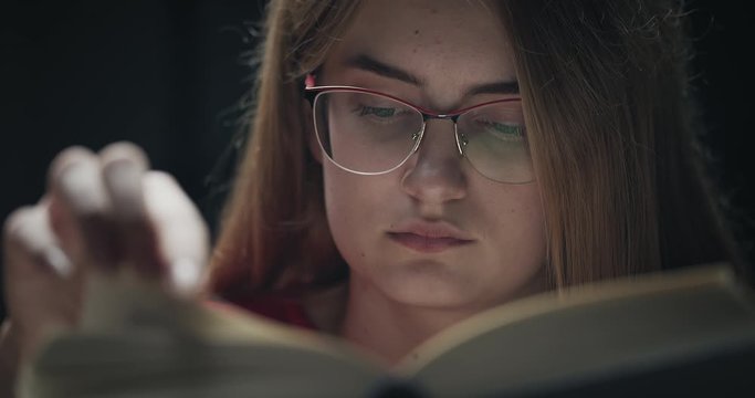 Young focused woman turning book page in dark room, reading novel, face closeup