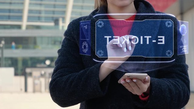 Unrecognizable woman interacts HUD hologram with text E-ticket. Girl in the coat uses the technology of the future mobile screen on the background of the city