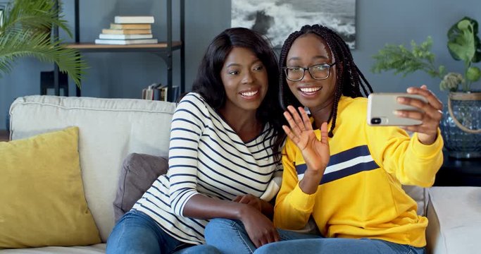 Two young joyful African American friendly girls smiling to the smartphone camera and waving with hands while taking selfie photos and sitting on the couch in the living room.