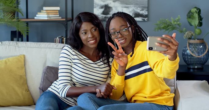 Two African American young happy girls, best friends laughing and waving with hands while posing and taking selfie photos on the smartphone camera on the sofa at home.
