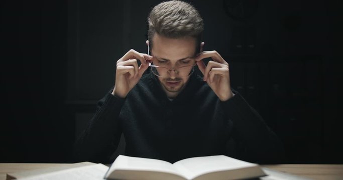 Tired male student taking off eyeglasses and rubbing nose bridge after reading books in darkness, overload