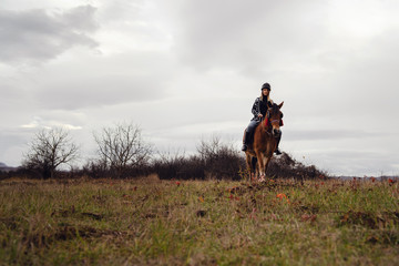 Young caucasian blonde woman female girl on the horse riding in nature wearing helmet in winter or autumn day against a a gray sky in the field