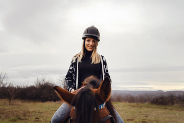 Young caucasian blonde woman female girl on the horse riding in nature wearing helmet in winter or...