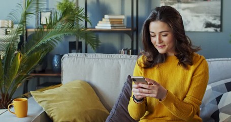 Young Caucasian attractive woman texting a message on the smartphone while chatting at home on the cozy couch. Inside. - Powered by Adobe