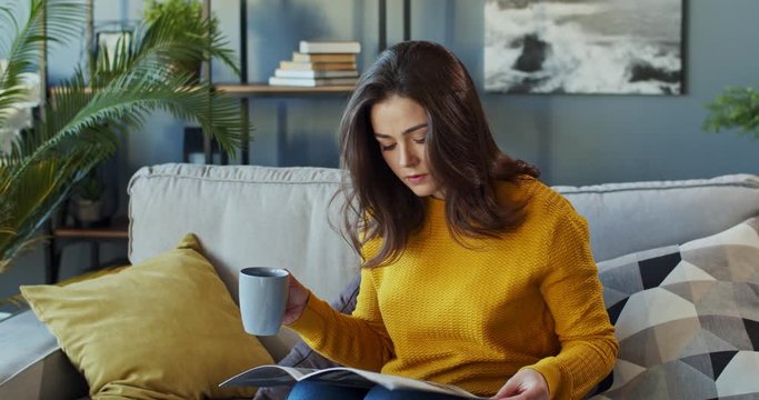 Caucasian Young Attractive Woman Sitting On The Couch In The Cozy Living Room, Sipping Hot Tea Or Coffee And Reading A Fashion Magazine. Indoors.