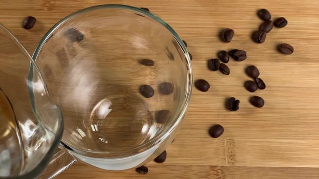 Top shot of pouring hot black coffee into a transparent glass cup. Coffee beans on the wooden table. Coffee foam closeup.