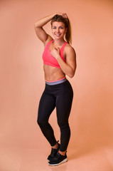 Fototapeta premium Vertical portrait of a pretty fitness blonde girl with a beautiful smile, excellent teeth and a bodybuilder figure in a tracksuit on a pink background. Cute looks at the camera, standing straight.
