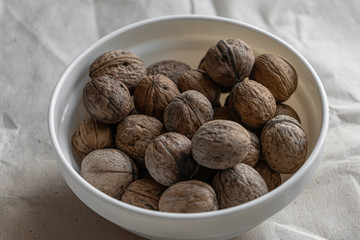 A white plate full of inshell walnuts stands on a brown background from crumpled old kraft paper. Top view flatlay
