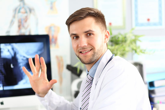 Portrait of smiling doc posing in hospital cabinet. Smiling surgeon wearing medical coat. X-ray picture of body on display of computer. Medicine treatment and healthcare concept. Blurred background