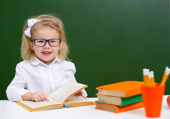 Cute little girl in glasses smiles cheerfully while reading a book in the classroom.