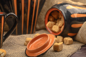 Pieces of brown sugar, a sugar bowl, and pottery against a background of coarse homespun cloth. Close up.