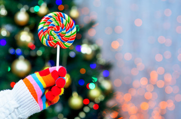 Fototapeta na wymiar hand in multi-colored glove holds a lollipop against the background of the Christmas tree
