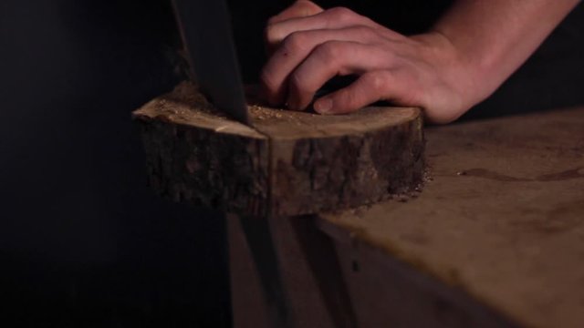 Close up of a carpenter using a saw to cut through a circular chunk of a tree trunk, with saw dust falling off the table top