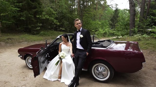 Beautiful young European married couple stay next to the old cherry colour Ford Mustang in the middle of the forest