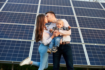 Young couple kisses with a baby in her arms on a background of solar panels. Blond kid is looking...